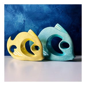 May include: Two ceramic fish sculptures. One is yellow, the other is light blue. Both have a large circular opening in the center and a smaller eye-shaped opening. The background is a textured dark blue.