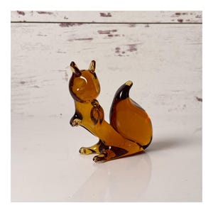 May include: A glass squirrel figurine in amber and dark brown. The squirrel is posed upright, with its tail curved upwards. The figurine is made of transparent glass, allowing light to pass through. The squirrel is a decorative item.