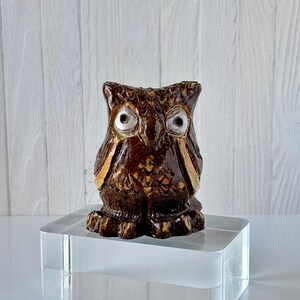 May include: A ceramic owl figurine with a glossy brown finish and gold accents. The owl has large, detailed eyes and is perched on a clear acrylic block. The background is a white wooden panel.