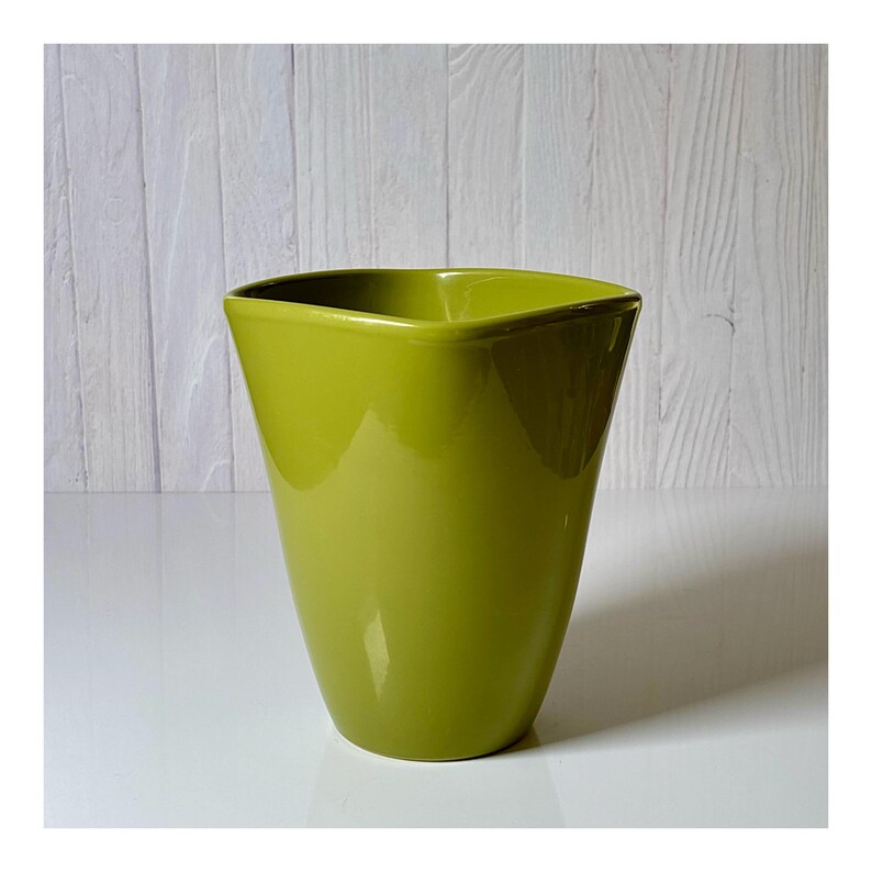 May include: A glossy, lime green ceramic vase with a unique, angular design. The vase has a square top that tapers down to a wider base. The smooth surface reflects light, highlighting its vibrant color and modern shape.