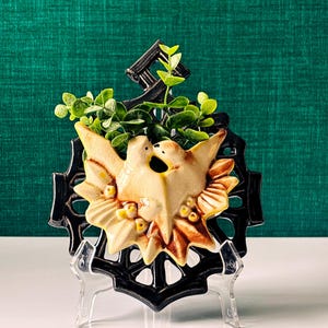 Peut inclure: Décoration murale en céramique avec deux colombes qui s'embrassent, dans des tons crème et marron, entourées d'un cadre noir décoratif. La pièce est ornée de petits accents jaunes et de feuillage vert, et est présentée sur un support transparent.