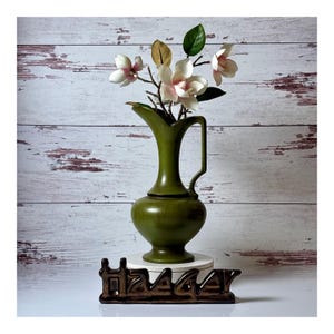 May include: An olive green ceramic pitcher with a slender neck, a curved handle, and a rounded base, containing a bouquet of white and pink magnolia flowers with green leaves, positioned above a dark brown decorative base with cutout lettering.