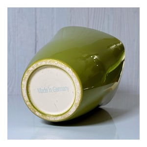 May include: A glossy, olive-green ceramic vase with a unique, asymmetrical shape. The base is off-white with the text "Made in Germany" in light blue. The vase has a smooth, reflective surface.
