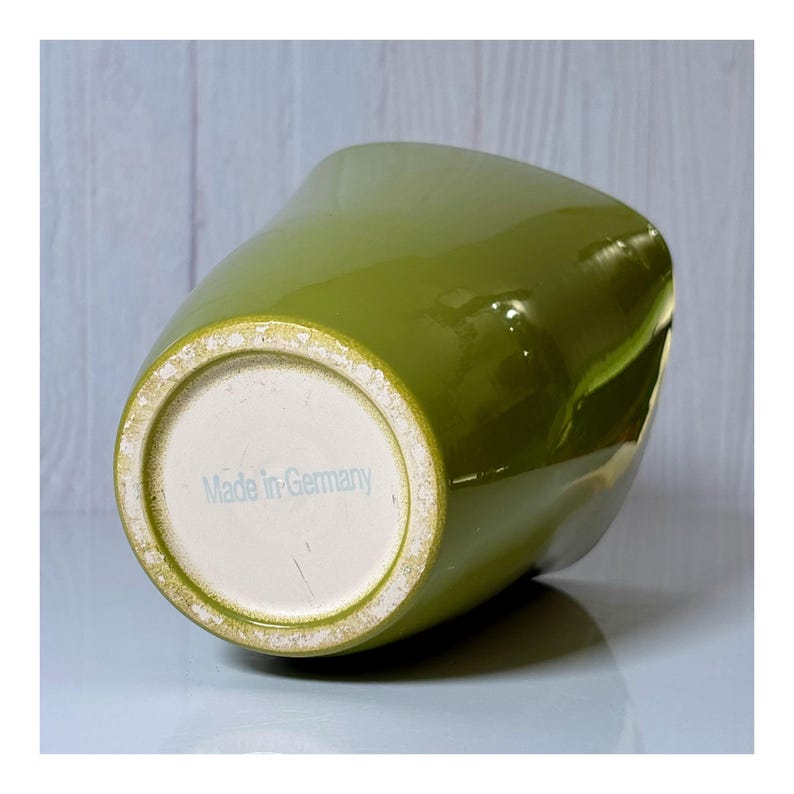 May include: A glossy, olive-green ceramic vase with a unique, asymmetrical shape. The base is off-white with the text "Made in Germany" in light blue. The vase has a smooth, reflective surface.