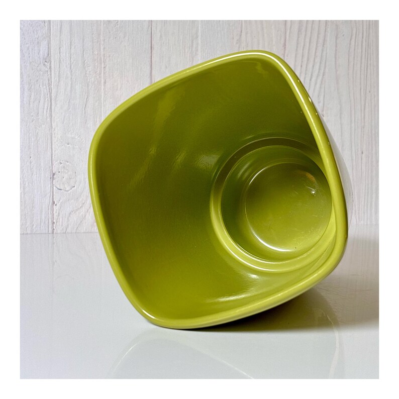 May include: A glossy, lime green ceramic planter with a square, rounded shape. The interior is a lighter shade of green. The planter sits on a white surface, with a white wood-paneled background.