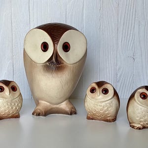 May include: A collection of four owl figurines. The largest owl is tall with a brown head and large, off-white eyes with red pupils. Three smaller owls have similar features. The figurines are primarily beige and brown.