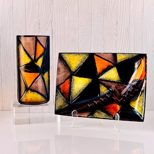 May include: Two decorative glass items with geometric patterns. A rectangular vase and a rectangular plate, both with yellow, orange, brown, and black triangles. The vase is on a clear acrylic stand. The plate is on a clear stand.