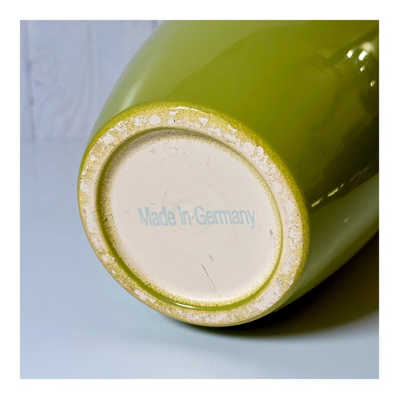 May include: A green ceramic vase with a wide base and a slightly tapered body. The bottom of the vase is off-white with the text "Made in Germany" in light blue. The vase has a glossy finish.