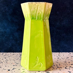 Puede incluir: Un jarrón hexagonal alto con un cuerpo verde brillante y un borde verde claro y beige. El jarrón de cerámica presenta diseños florales en relieve y es adecuado para exhibir flores o como pieza decorativa.