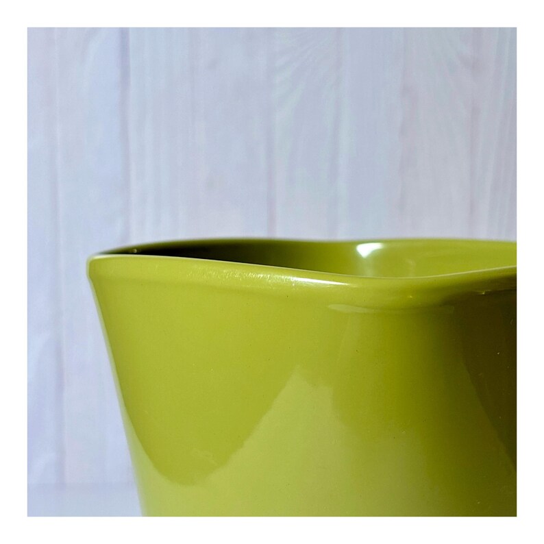 May include: Close-up of a lime green ceramic pitcher with a wavy rim. The pitcher has a glossy finish and a smooth surface. The background is a light-colored wooden surface.