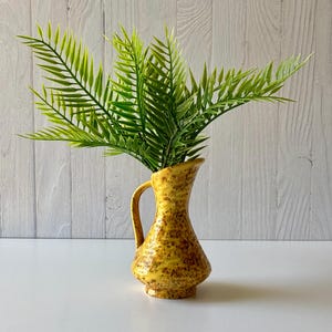 May include: A yellow ceramic jug with a speckled pattern, holding vibrant green fern-like leaves. The jug has a handle and a flared top. The background is a light-coloured wooden surface.