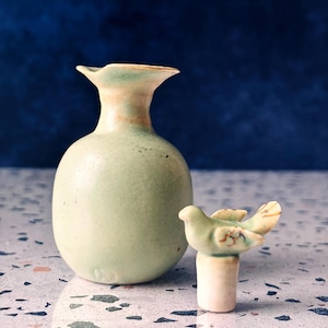 May include: A small, pale green ceramic vase with a rounded body and flared rim, next to a small bird figurine. The bird is perched on a cylindrical base, with green and brown details. The background is a dark blue.