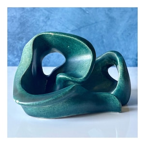 Può includere: Una scultura in ceramica color verde ottanio scuro con una finitura liscia e lucida e una forma astratta e fluida. L'oggetto presenta molteplici anelli curvi a forma di nastro e spazi aperti, creando una struttura continua e interconnessa.