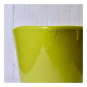 May include: A close-up of a lime green ceramic planter. The planter has a glossy finish and a slightly tapered shape. The background is a light-colored wooden surface.
