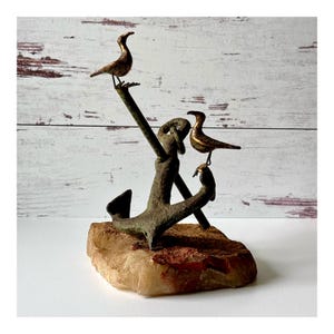 May include: A decorative sculpture featuring two metallic gold-toned birds perched on a dark, textured metal anchor. The anchor is mounted on a rough, light-colored stone base with reddish-brown mineral deposits, creating a nautical-themed display.