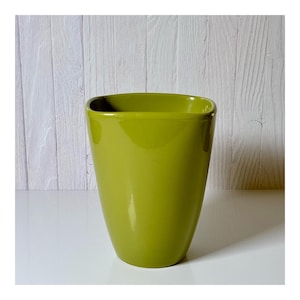 May include: A glossy, olive-green ceramic planter with a square top and rounded corners. The planter has a smooth, tapered design and is suitable for indoor or outdoor use. The background is a white wooden surface.