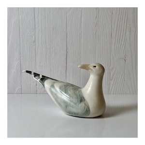 May include: A ceramic seagull figurine in a resting pose. The bird has a white body with gray and black markings on the wings and tail. The beak is a light yellow color. The figurine is on a white surface with a light wood-paneled background.