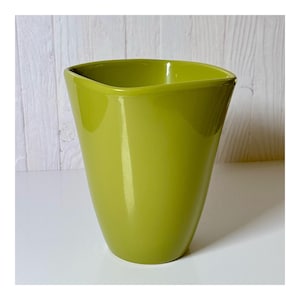 May include: A glossy, lime green ceramic vase with a unique, asymmetrical top. The vase has a smooth, curved shape and a wide opening. The background is a white surface and a light wood-paneled wall.