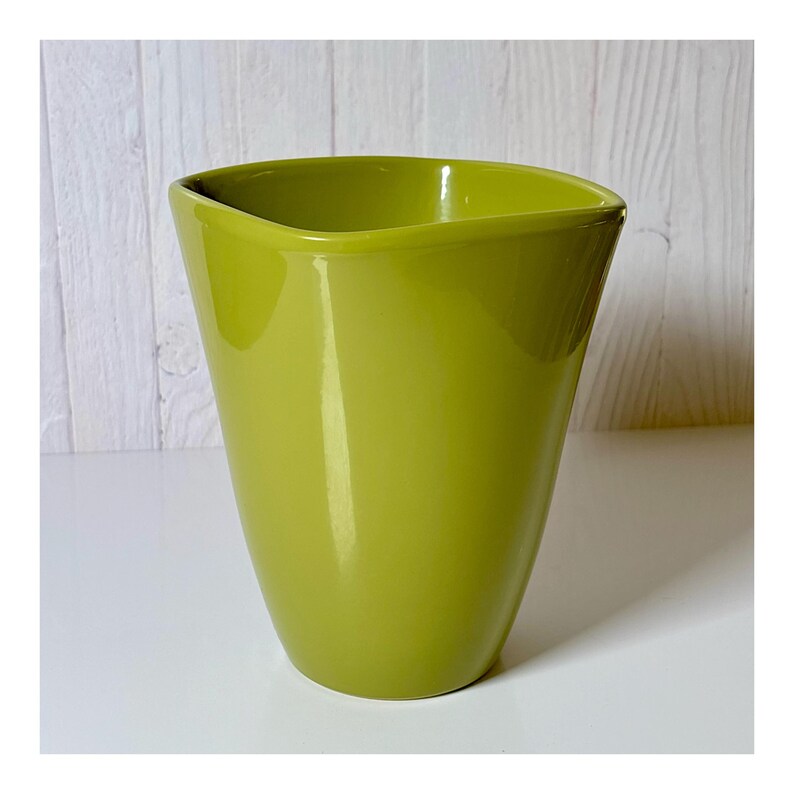 May include: A glossy, lime green ceramic vase with a unique, asymmetrical top. The vase has a smooth, curved shape and a wide opening. The background is a white surface and a light wood-paneled wall.