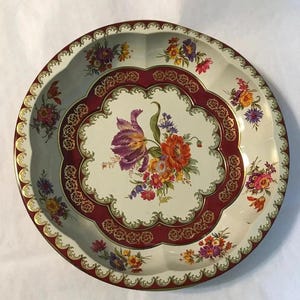 Puede incluir: Bandeja redonda de metal con fondo crema y diseño floral. La bandeja presenta un borde burdeos con detalles dorados y un arreglo floral central de flores moradas, naranjas y amarillas.