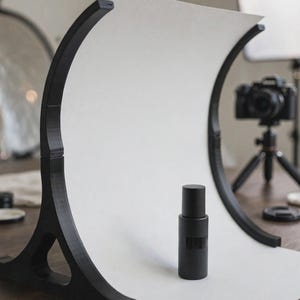 May include: A black matte cylindrical bottle stands on a white curved backdrop supported by a black 3D-printed frame. The frame features two arched side supports with triangular bases that hold the white paper in a smooth, continuous sweep.