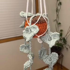 May include: A handmade crochet plant hanger with a pot. The pot is orange and brown, and the hanger is white with light blue leaf accents. The plant hanger is suspended from white cords.