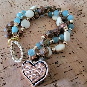 May include: A multi-strand beaded bracelet with a heart-shaped pendant. The bracelet features blue, brown, and white beads, with gold-colored metal accents. The heart pendant is encrusted with small, sparkling crystals. A woven loop is attached.