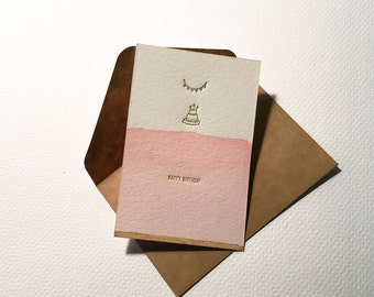 May include: A pink and white birthday card with gold foil accents. The card features a cake and a string of bunting. The text "Happy Birthday" is printed in gold foil.