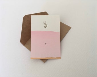 May include: A white card with a pink watercolour background and a simple illustration of a stocking. The text "JOY" is printed in black at the bottom of the card.