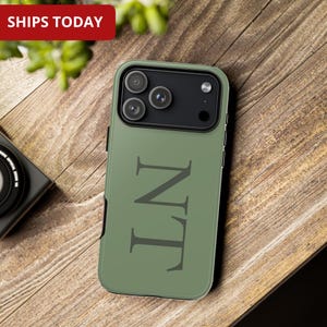 Puede incluir: Funda de teléfono verde oliva con las letras "NT" en gris oscuro. La funda tiene un módulo de cámara negro y un borde negro. El texto "SHIPS TODAY" está en un rectángulo rojo en la esquina superior izquierda.