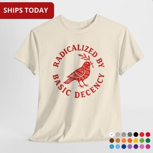 Puede incluir: Camiseta color crema con un gráfico rojo. El gráfico presenta un pájaro sosteniendo una rama, rodeado por las palabras "RADICALIZED BY BASIC DECENCY". La camiseta tiene mangas cortas y cuello redondo.