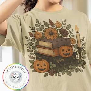 Puede incluir: Camiseta beige con un diseño de temática otoñal. El diseño incluye libros apilados, calabazas, flores y una vela. El texto "MULTIPLE COLOR OPTIONS AVAILABLE" está en un logotipo circular.