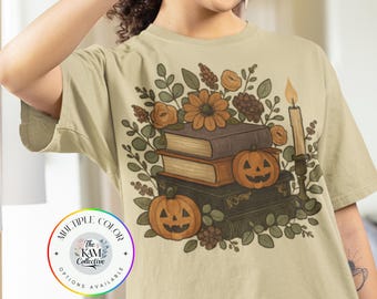 May include: Beige t-shirt featuring a fall-themed graphic. The design includes stacked books, pumpkins, flowers, and a candle. The text "MULTIPLE COLOR OPTIONS AVAILABLE" is in a circular logo.