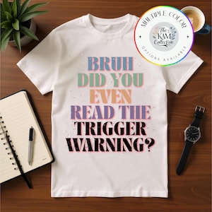 Può includere: T-shirt bianco con la scritta "BRUH DID YOU EVEN READ THE TRIGGER WARNING?" in lettere colorate e consumate. La maglietta presenta anche un logo circolare con la scritta "MULTIPLE COLOR OPTIONS AVAILABLE".