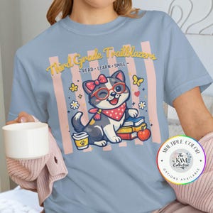 Puede incluir: Camiseta azul claro con un gato de dibujos animados con gafas y un pañuelo. El texto dice "Third Grade Trailblazers - READ. LEARN. SMILE." El diseño incluye libros, flores y una taza de café.