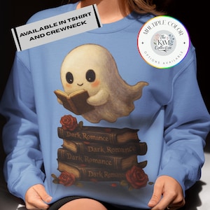 May include: Light blue sweatshirt featuring a cute ghost reading a book above a stack of books labeled "Dark Romance" with red roses. The text "AVAILABLE IN TSHIRT AND CREWNECK" is at the top. The KAM Collective logo is on the right.