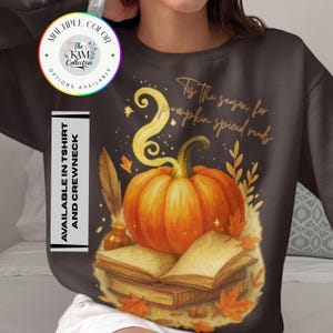 May include: Dark brown sweatshirt featuring a vibrant orange pumpkin atop a stack of books, with autumn leaves and a feather quill. Text reads "Tis the season for pumpkin spiced reads." Available in t-shirt and crewneck.