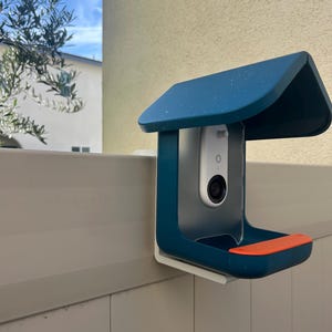 May include: A dark teal protective housing with a roof-like top and an orange accent piece mounted on a light beige fence. The housing contains a white vertical security camera with a black lens and a small circular button on its front surface.