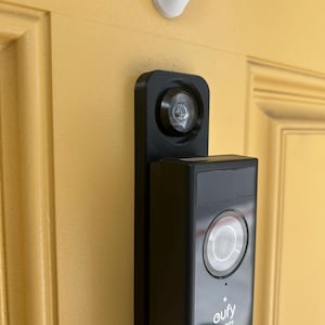 May include: A black Eufy Security video doorbell mounted on a yellow door. The doorbell has a camera lens and a button. A white hook is above the doorbell.