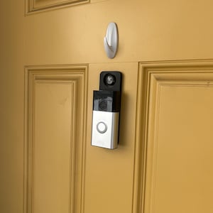 May include: A silver and black video doorbell with a white button and the brand name "ring" printed on the front. The doorbell is mounted on a yellow door next to a white hook.