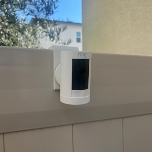 May include: A white cylindrical security camera with the word ring printed on the top front. The device features a black rectangular lens area and a perforated speaker section at the bottom, mounted securely onto the top edge of a beige fence panel.