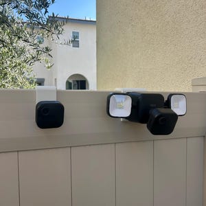 May include: A black Blink security camera and a black Blink floodlight camera system, both mounted on white brackets attached to a beige fence. The floodlight unit features two square LED light panels flanking a central camera lens.
