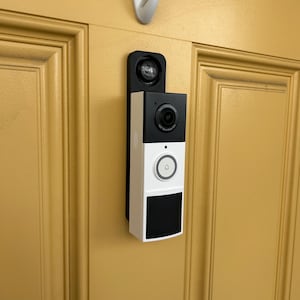 May include: A black and white smart doorbell with a camera and a button, mounted on a yellow door. The doorbell has a sleek, modern design and includes a small camera lens and a doorbell button. A white hook is mounted above the doorbell.