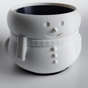 May include: A white snowman-shaped container with a black interior. The container has a smiling face, a scarf, and three buttons. The base is black.