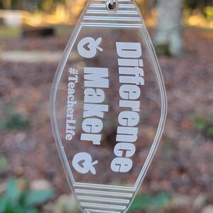 May include: Clear acrylic keychain with the words "Difference Maker" and "#TeacherLife" in white. The keychain has a silver chain and key ring. The background is a blurred view of trees.