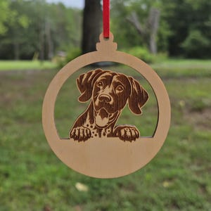 Può includere: Ornamento natalizio in legno con un design tagliato al laser di un cane. La testa e le zampe del cane sono visibili, che sbucano da un bordo. L'ornamento è circolare e ha un nastro rosso per appenderlo.