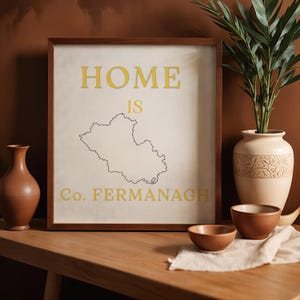 May include: A framed print with the words "HOME IS" above a map outline and "Co. FERMANAGH" in yellow. The print is in a brown wooden frame, displayed on a wooden table with decorative vases and bowls.