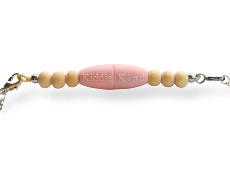 May include: A baby pink pacifier clip with the text "Kiddie Kind" in the center. The clip features wooden beads and a silver chain. The clip is designed to attach to a pacifier.