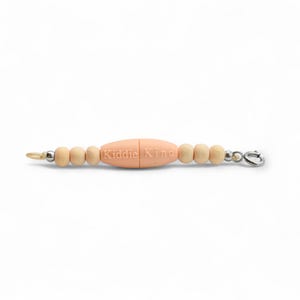 May include: A baby pacifier clip with a central peach-colored bead engraved with "Kiddie King". The clip features wooden beads and a metal clasp. The clip is designed to attach to a pacifier or toy.