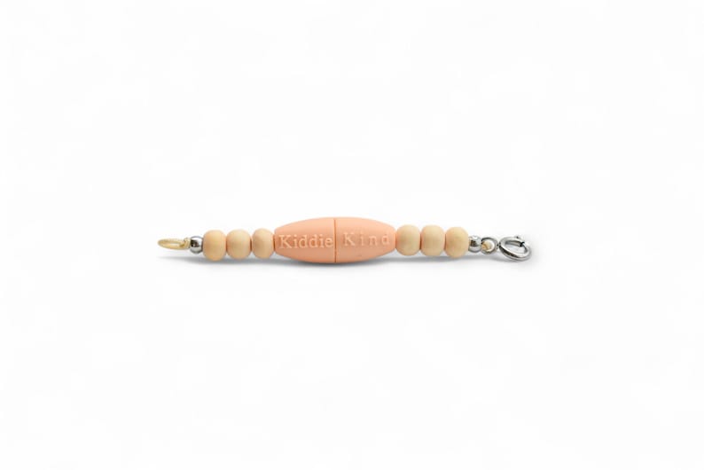 May include: A baby pacifier clip with a central peach-colored bead engraved with "Kiddie King". The clip features wooden beads and a metal clasp. The clip is designed to attach to a pacifier or toy.